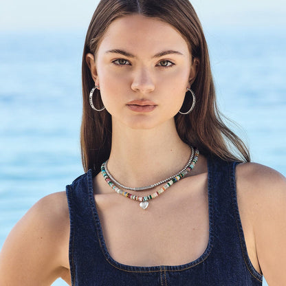 Model wearing a Brighton beaded necklace with multicolored stones and silver-plated heart pendant, paired with hoop earrings