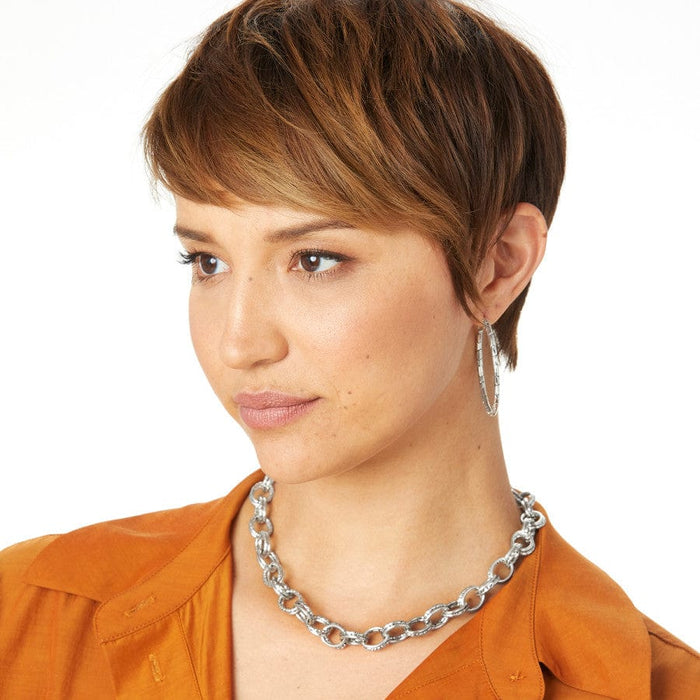A model wearing Pebble Large Hoop Earrings with short hair while glancing away with a matching silver necklace and orange top, against a light background.